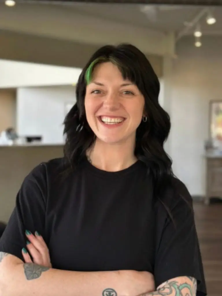 Smiling woman with black hair and a green streak, wearing a black T‑shirt with arms crossed; visible forearm tattoos, indoors.