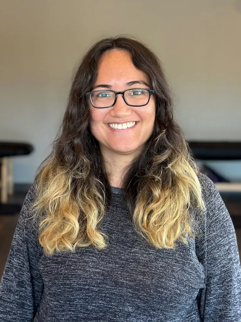 Woman with long wavy hair and glasses, wearing a dark gray sweater, smiling at the camera.