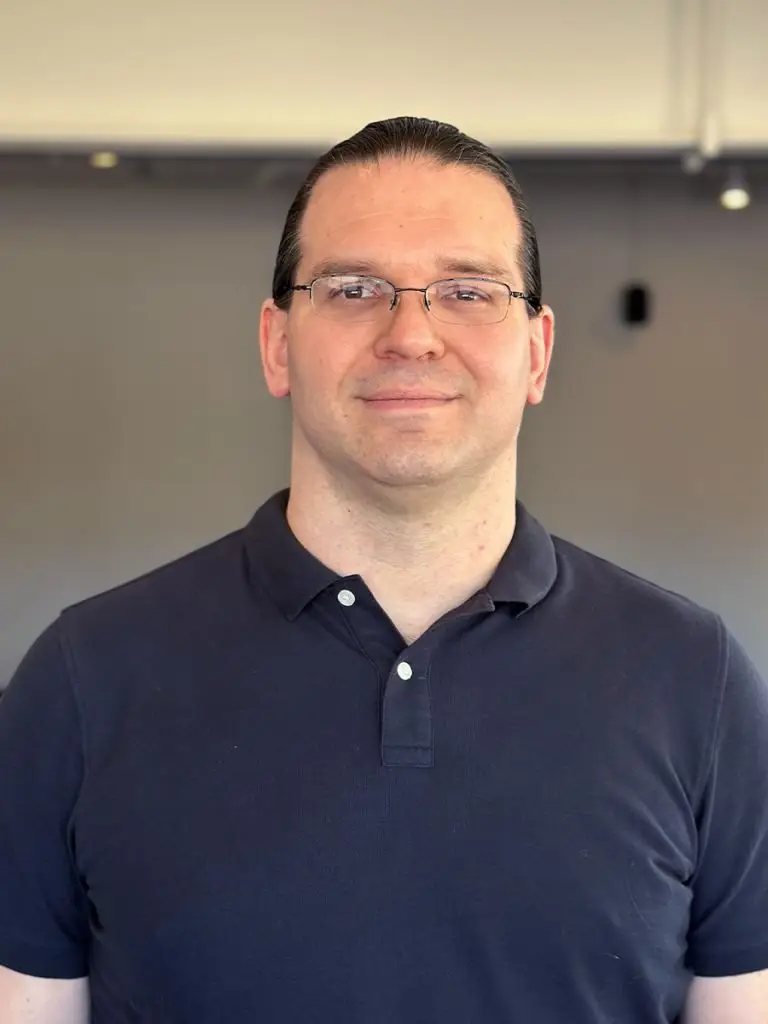 Head-and-shoulders portrait of a man with glasses wearing a navy polo shirt, neutral background.